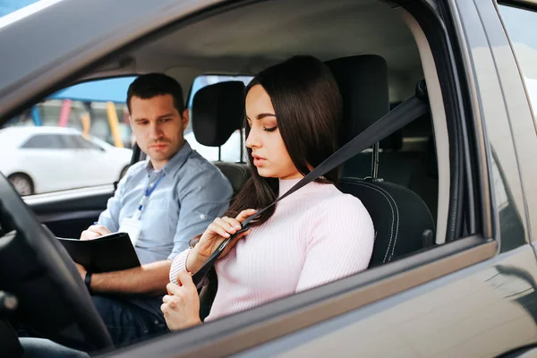 Homme instructeur auto prend examen chez la jeune femme. Brunette tenir la main sur la ceinture de sécurité et le verrouiller. Jeune homme assis en outre avec des papiers d'examen . — Photo