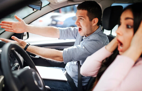 Male auto instructor takes exam in young woman. Stress during driving. Scared young woman touch head with hands. Instructor screaming and pointing forward.