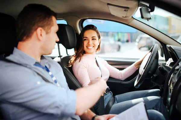 Hombre auto instructor toma examen en mujer joven. Morena mira al chico y sonríe. Conducir el coche con confianza. Punto de hombre con mano . — Foto de Stock