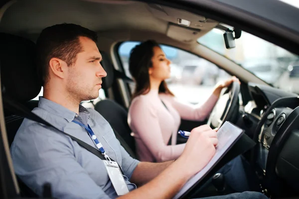 Male auto Instructor neemt examen in jonge vrouw. Man kijk uit en schrijf. Jonge vrouw rijden auto voorzichtig en met aandacht. — Stockfoto