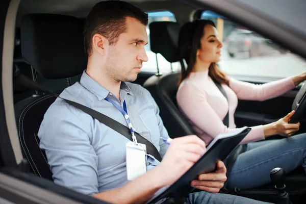 Homme instructeur auto prend examen chez la jeune femme. Attention regard sérieux tout droit sur la route. Ecris sur papier. Jeune femme conduite prudente. Concentré. Réussir le test pratique . — Photo