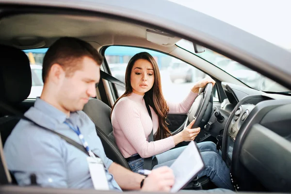 Male auto instructor takes exam in young woman. Serious concentrated guy writing on paper. Wondered woman look at it. Holding hands on steering wheel. Passing practical test. — 스톡 사진