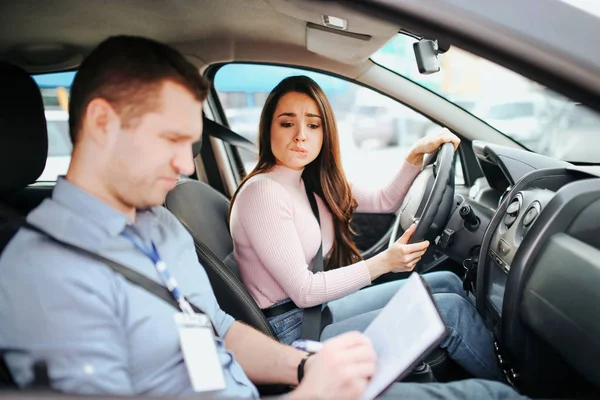 Hombre auto instructor toma examen en mujer joven. Mujer joven preocupada mirar los resultados de las pruebas. Tomando las manos en el volante. El hombre confundido mira hacia abajo. Luz del día . — Foto de Stock