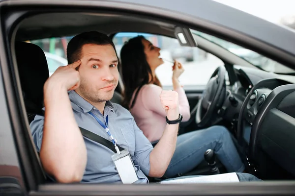 Homme instructeur auto prend examen chez la jeune femme. Regardez la caméra et pointez la tête avec le doigt. Étudiant fou se maquillant en voiture. Regardez sur le miroir . — Photo