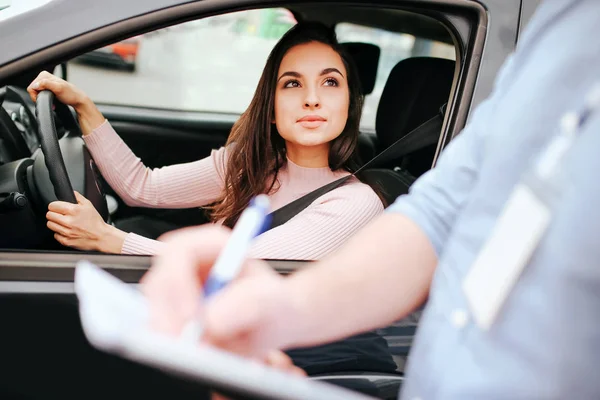Homme instructeur auto prend examen chez la jeune femme. Il écrit le résultat du test dans du papier. Femme intéressante regarder gars dehors de la voiture. Tenez les mains sur le volant et la ceinture de sécurité à travers le corps . — Photo