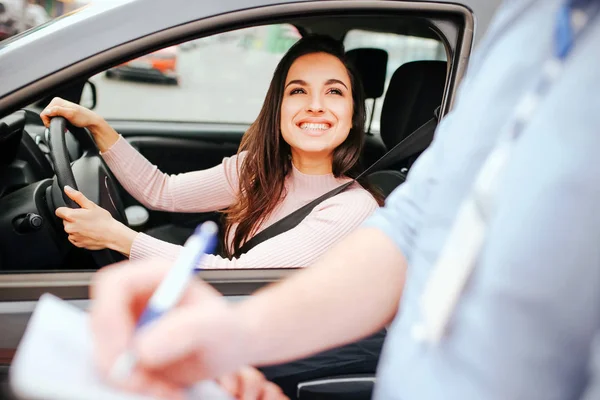 Maschio istruttore di auto prende esame in giovane donna. Allegro modello attraente guardare ragazzo e sorridere. Tenendo le mani sul volante. Marchio di scrittura dell'istruttore su carta . — Foto Stock