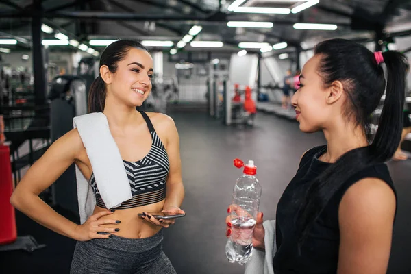 Fröhliche und sexy junge Frau lächelt ihrem asiatischen Freund zu. sie schaut sie an und hält die Hände an den Hüften. weißes Handtuch liegt auf der Schulter. Auch asiatische Models lächeln. sie hat eine Wasserflasche. — Stockfoto