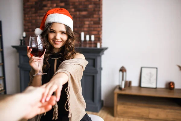 Joven hermosa mujer atractiva en sombrero rojo posando en la cámara y sonreír. Celebrando año nuevo o Navidad. Sostenga la copa de vino tinto y mans mano. Luz del día . — Foto de Stock