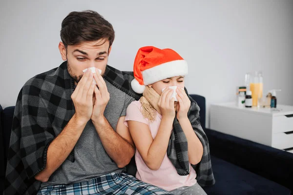 Picture of young man with daughter sitting on sofa and sneezing. They caught cold. Flu and sickness. Celebrating new year of Christmas. Red hat of girls head. Festive month. — Stock Photo, Image