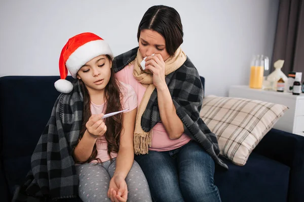 Sick young woman hold white tissue close to nose and look at thermomenter. Girl hold it and look too. Wear festive red hat. Christmas or new year period. Cozy warm blanket on shoulders.