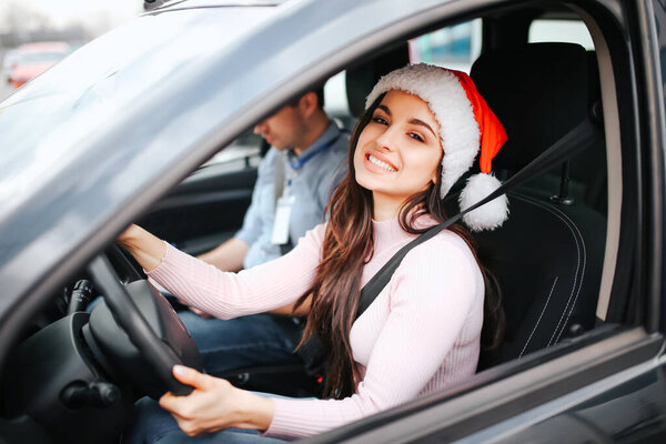 Picture of attractive young woman sit on drivers place. Hold hands on steering wheel and smile. Christmas or new year time. Wear red hat and smile. Man sit beside.