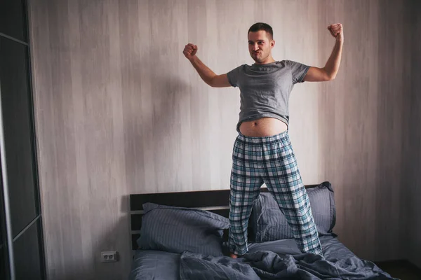 Un hombre con panza se para en la cama y hace muecas. Frick, concepto divertido casa modelo masculino — Foto de Stock