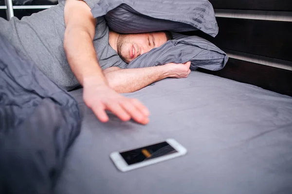 Não há força para sair da cama de manhã. Um homem de manhã na cama não quer acordar. quer desligar o alarme . — Fotografia de Stock