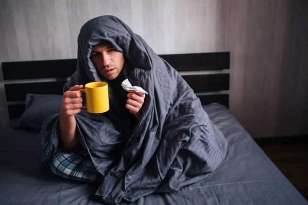 He covered himself with a blanket and holds a napkin and hot tea. The man got sick. Lies in bed and suffers. He has fever, flu, colds and snot.