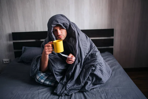 El hombre se enfermó. Se acuesta en la cama y sufre. Tiene fiebre, gripe, resfriados y mocos. Se cubrió con una manta y sostiene una servilleta y té caliente. . Imagen De Stock