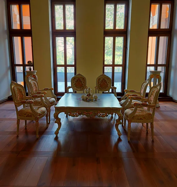 A square table on brown parquet with tea-set and six white-cream elegant chairs in a classic interior. Vintage style. Big glass windows.