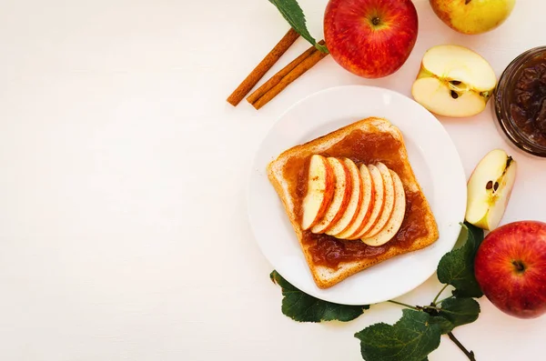 Toast mit zerdrückter Apfelmarmelade, Scheiben frischer Äpfel, Zimt auf einem Teller auf einem weißen Tisch. Äpfel und Apfelmarmelade im Glas. das Konzept. leckeres süßes Frühstück. Ansicht von oben. — Stockfoto