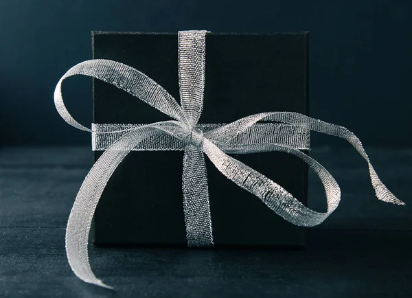 Black gift box with silver ribbon bow on black background. Tonned photo.