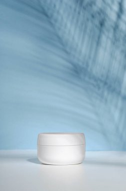 A white jar of cream on a stand on a blue background, on a white table with tropical palm leaves and their shadow. Stylish look of the product, mock up, identity clipart