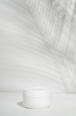 Beyaz arka planda beyaz bir krema kavanozu ve tropikal palmiye yapraklarının gölgesi. Ürünün şık görünümü, modeli, kimliği.