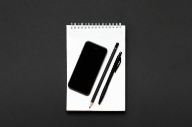Siyah arka planda siyah kalem, kalem ve akıllı telefon olan beyaz defter. Ofis masası, kırtasiye. Üst manzara, düz uzanma