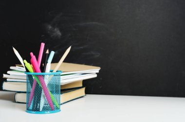 Çok renkli kalemler, stantta kalemler ve beyaz bir masaya dizilmiş defterli kitaplar tebeşirle boyanmış siyah bir okulun kara tahtasının arka planına karşı. Boşluğu kopyala