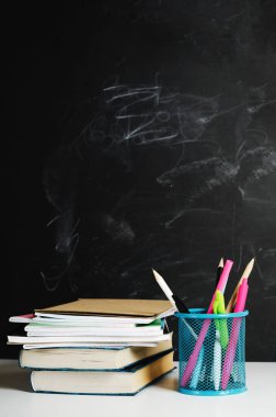Çok renkli kalemler, stantta kalemler ve beyaz bir masaya dizilmiş defterli kitaplar tebeşirle boyanmış siyah bir okulun kara tahtasının arka planına karşı. Boşluğu kopyala