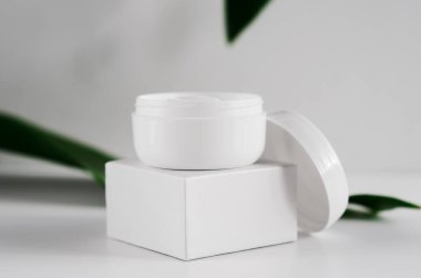 Doğal kozmetik krem kavanozu ve yeşillik manzarası. Arkaplanda beyaz plastik kaplar ve bulanık bitki yaprakları var. Organik kozmetoloji yaratıcı kavramı. Cilt bakımı, hijyen aksesuarı.