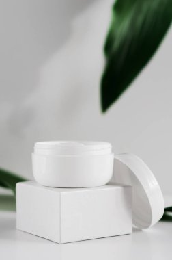 Doğal kozmetik krem kavanozu ve yeşillik manzarası. Arkaplanda beyaz plastik kaplar ve bulanık bitki yaprakları var. Organik kozmetoloji yaratıcı kavramı. Cilt bakımı, hijyen aksesuarı.