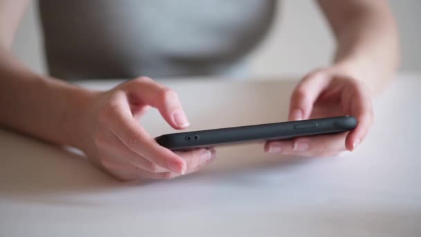 Una joven se sienta en su casa en una mesa y sostiene un dispositivo móvil en sus manos. La generación actual de millennials utiliza sus teléfonos en casa. Jugar en un smartphone móvil — Vídeo de stock