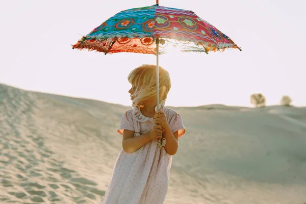 Little lonely girl with bright umbrella  walking on a dune in the desert