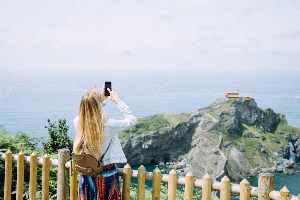 Side view of a young tourist woman taking picture of a mirador de san juan de gaztelugatxe
