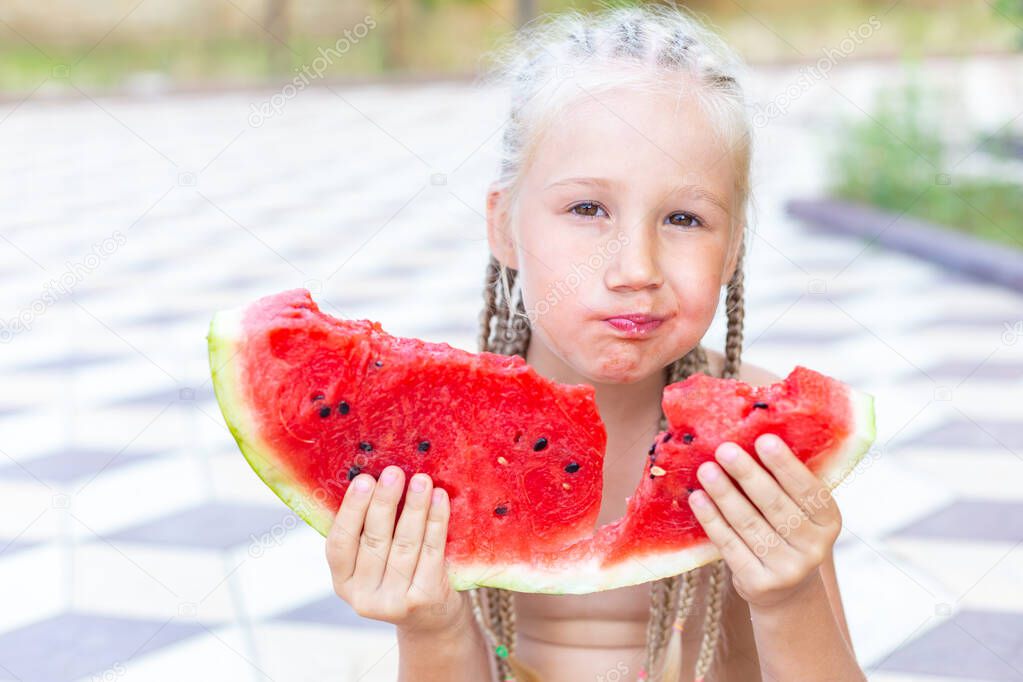 Beautiful caucasian little girl with long blond pigtails holds a juicy ripe red slice of watermelon in her hands, looks at the camera, chews, smiles. National Watermelon Day. Portrait waist-high.