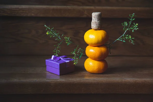 Tangerine snowman, green twigs and champagne corks. Gift, purple box. Wooden background, staircase. New Year, Christmas, traditions. A creative approach to gift design.