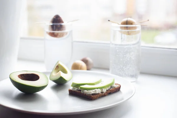 Sliced avocado and avocado sandwich on a plate by the window. A avocado seed sprouts in a glass of water. Healthy lifestyle concept. Houseplants.