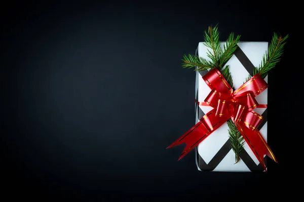 Regalo di Natale splendidamente avvolto in carta bianco-nera, fiocco rosso, ramo di abete rosso, sfondo nero.. — Foto Stock