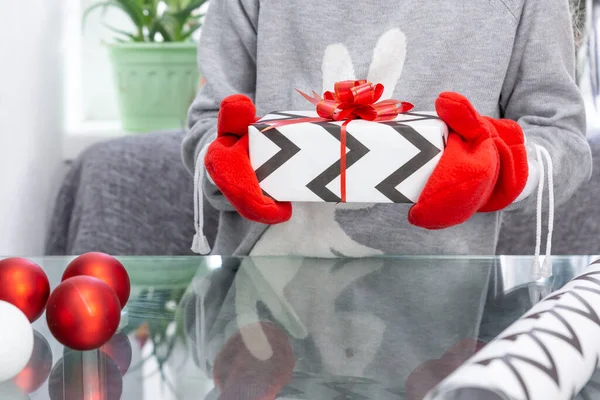 Una niña en un suéter gris con un conejo sostiene un regalo con manoplas rojas en las manos. Año Nuevo, Navidad, vacaciones de invierno — Foto de Stock