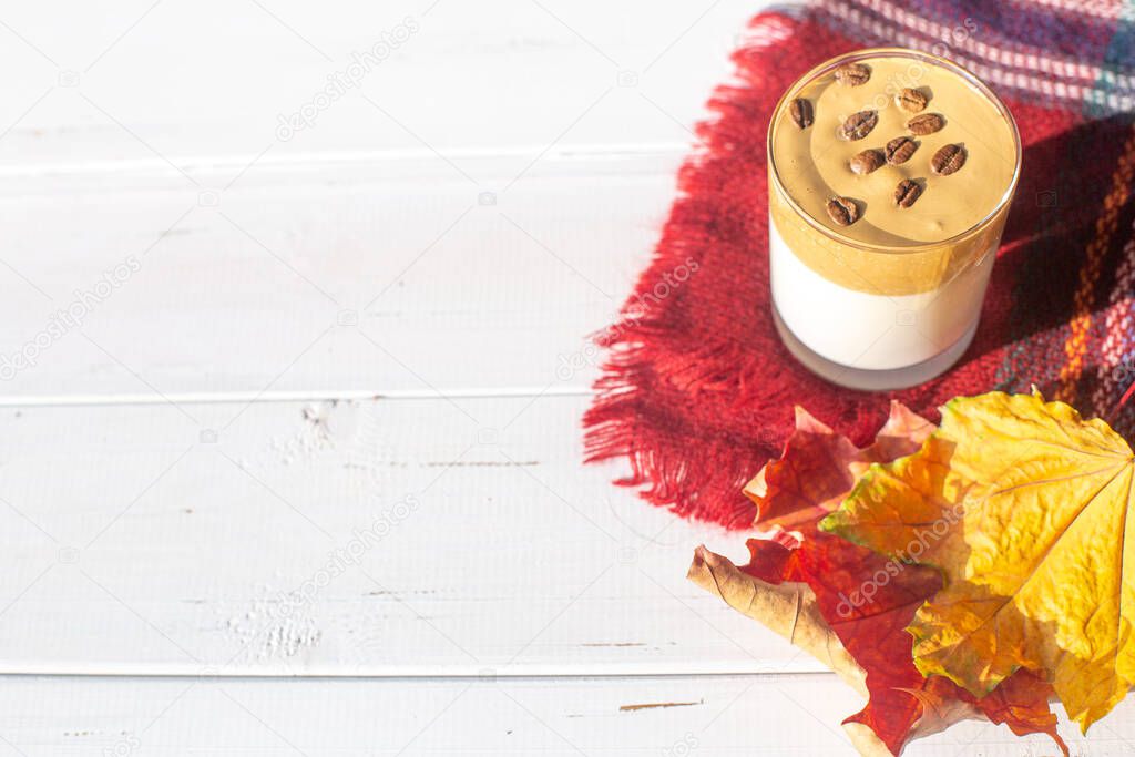 Traditional Korean caffeinated milk drink, Dalgona coffee in a glass with coffee beans on a white board background. Dry maple leaves. Warm checkered scarf or plaid. Autumn breakfast in cafe.Copy space