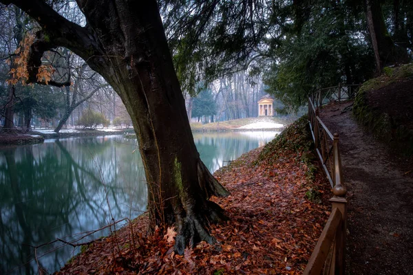 Little neoclassical building reflected onto a frozen lake surrounded by snow, Park of Monza, Italy