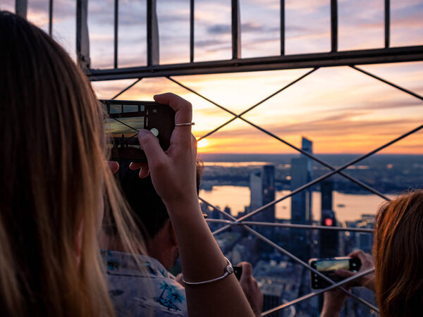 Young girls using their phones and cameras to take images of the city skyline from the observation platform of the Empire State Building, New York, USA