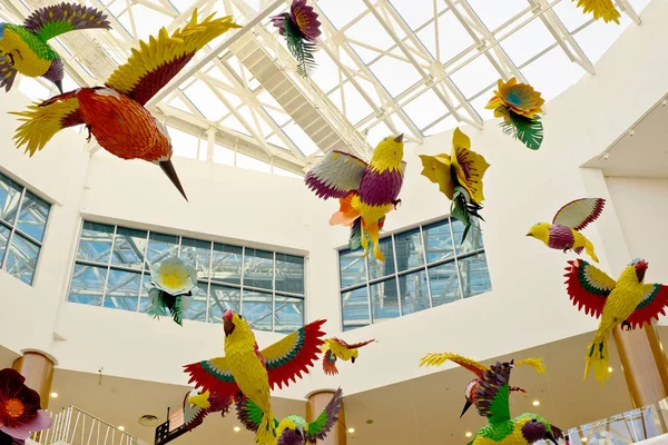 Collected from colored paper and glue, a flock of birds and flowers under the dome of the shopping center hang on thin transparent ropes. Right overhead.