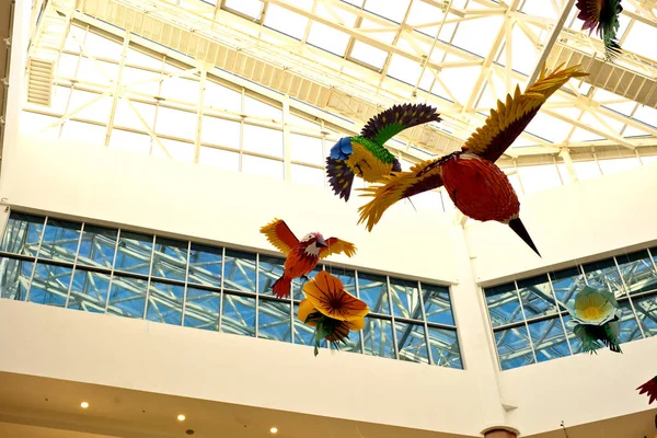 Collected from colored paper and glue, a flock of birds and flowers under the dome of the shopping center hang on thin transparent ropes. Right overhead.