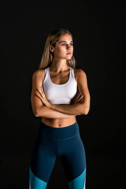 Siyah arka planda fitness eğitimi için spor üniformalı sarışın bir kızın yarım boy portresi. Kollar önünüzde göğüs hizasında katlandı.