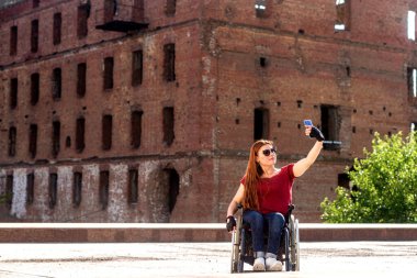 Engelliler için ulaşılabilir çevre kavramı. Tekerlekli sandalyedeki bir kız, tarihi bir anıtın arka planında kendi fotoğrafını çeker (İkinci Dünya Savaşı 'ndan kalma yarı yıkık bir ev))
