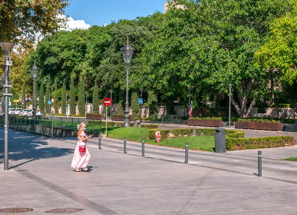 Palma de Mallorca, Balearic Adaları / İspanya; Eylül 2020: akıllı telefonuna ve maskesine bakan açık renk elbiseli kadın, Palma de Mallorca 'da güneşli bir caddede tek başına yürüyor.