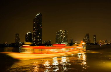Bu fotoğraf gece suda hızla hareket eden bir tekneyi yakalar. Teknenin ışıkları, başkentteki sahil şeridi boyunca uzanan gökdelenlerin ve binaların parlak ışıklarıyla çelişen güzel, bulanık bir renk çizgisi oluşturuyor.