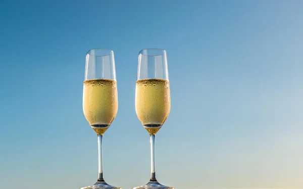 Golden champagne bubbles in flute glasses. Side view of two glasses of sparkling white wine isolated on blue sky background.