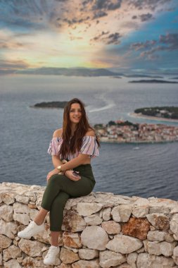 Sıcak bir yaz gününde taş bir duvarda oturan çekici esmer. Arka planda Adriyatik Denizi ve küçük Primosten kasabasıyla poz veren modaya uygun bir kız. Güzel altın gün batımı