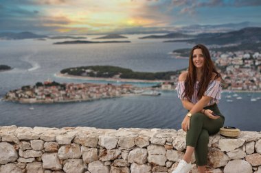 Sıcak bir yaz gününde taş bir duvarda oturan çekici esmer. Arka planda Adriyatik Denizi ve küçük Primosten kasabasıyla poz veren modaya uygun bir kız. Güzel altın gün batımı