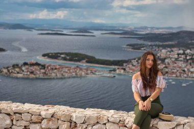 Sıcak bir yaz gününde taş bir duvarda oturan çekici esmer. Arka planda Adriyatik Denizi ve küçük Primosten kasabasıyla poz veren modaya uygun bir kız.